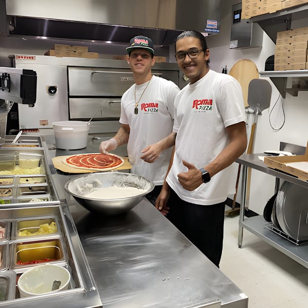 Two smiling Roma Pizza chefs in a commercial kitchen, one preparing a pizza with sauce.