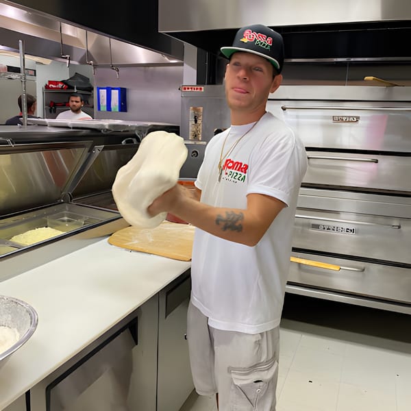 A Roma Pizza chef in a black cap and white shirt expertly tosses pizza dough in a kitchen.